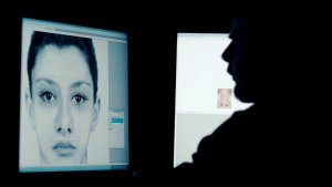Zrzut z ekranu. Na ekranie monitora szkic twarzy kobiety. Przed monitorem siedzący mężczyzna. Pomieszczenie wyciemnione.