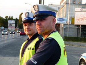 Zdjęcie kolorowe. Dwóch policjantów ruchu drogowego w umundurowaniu służbowym i kamizelkach odblaskowych nadzorujących ruch na drodze.