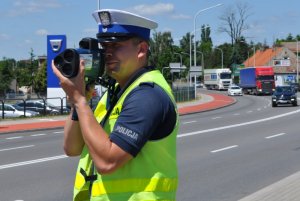 Zdjęcie kolorowe. Policjant ruchu drogowego mierzy prędkość przejeżdżających pojazdów.