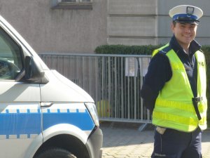 Zdjęcie kolorowe. Policjant ruchu drogowego w umundurowaniu służbowym i kamizelce odblaskowej podczas służby na drodze. Z prawej strony fragment oznakowanego radiowozu Policji.