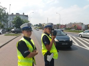 Zdjęcie kolorowe. Policjantki ruchu drogowego w umundurowaniu służbowym i odblaskowych kamizelkach, kontrolują prędkość przy drodze.