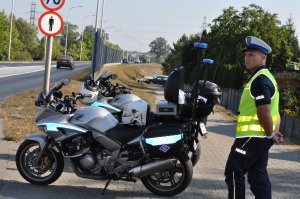 Zdjęcie kolorowe. Policjant ruchu drogowego w umundurowaniu służbowym, stojący obok oznakowanego, policyjnego motocykla.