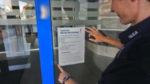 na zdjęciu policjantka w umundurowaniu służbowych na drzwiach wejściowych do banku umieszcza plakat Seniorze nie daj się oszukać
