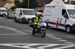 Zdjęcie kolorowe. Policjant ruchu drogowego na oznakowanym motocyklu podczas zabezpieczenia przemarszu pielgrzymki. W tle samochody.