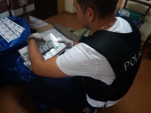 Zdjęcie kolorowe. Policjant w kamizelce taktycznej z napisem policja pochylający się nad zabezpieczonymi kartonami z paczkami papierosów.