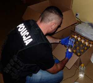 Zdjęcie kolorowe. Nieumundurowany policjant, w kamizelce taktycznej z napisem Policja trzyma w dłoniach zabezpieczona butelkę z nielegalnym spirytusem.