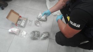 Zdjęcie kolorowe.  Rozłożone na podłodze woreczki foliowe z zabezpieczonymi narkotykami. Obok umundurowany policjant.
