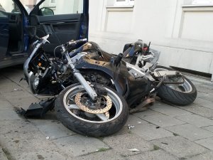 Zdjęcie kolorowe. Motocykl koloru czarno-srebrnego leżący na chodniku. Wokół odłamki uszkodzonych elementów pojazdów.
