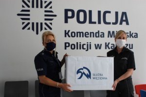 zdjęcie kolorowe na zdjęciu funkcjonariuszka policji oraz służby więziennej przekazująca maseczki ochronne uszyte przez skazanych