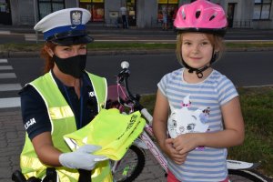 Zdjęcie kolorowe. Policjantka ruchu drogowego w umundurowaniu służbowym i kamizelce odblaskowej przekazuje kamizelkę odblaskową dziewczynce - rowerzystce).