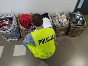 Zdjęcie kolorowe. Policjant  w odblaskowej kamizelce z napisem &quot;Policja&quot; prowadzi oględziny zabezpieczonej odzieży z podrobionymi znakami towarowymi. Odzież znajduje się w pięciu torbach.