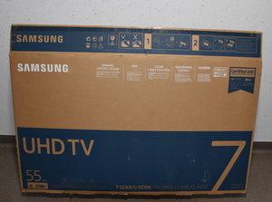 Zdjęcie kolorowe. Karton stojący na podłodze. Na kartonie duże napisy Samsung, UHD TV 55.