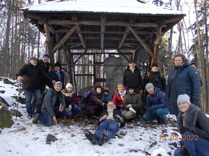Zdjęcie kolorowe.Grupa osób pozujących do zdjęcia na tle kamiennych posągów umieszczonych pod drewnianym zadaszeniem otoczonych metalową siatką. W tle las.