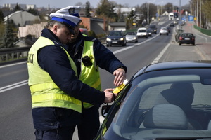 Zdjęcie kolorowe. Policjanci z ruchu drogowego w umundurowaniu służbowym i kamizelkach odblaskowych, przeprowadzają badanie stanu trzeźwości zatrzymanego do kontroli drogowej kierowcy.