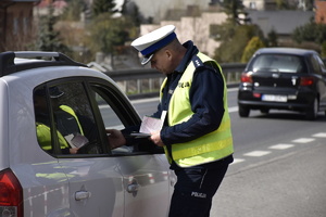 Zdjęcie kolorowe. Policjant z ruchu drogowego w umundurowaniu służbowym i kamizelce odblaskowej, wręcza kierowcy zatrzymanemu do kontroli mandat karny.