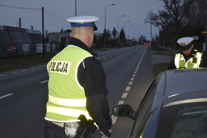 Zdjęcie kolorowe. Policjanci z ruchu drogowego w umundurowaniu służbowym i kamizelkach odblaskowych, przeprowadzają kontrolę drogową zatrzymanego kierowcy.