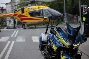 Zdjęcie kolorowe. Na pierwszym planie po prawej stronie fotografii - policyjny motocykl. W tle na środku drogi helikopter Lotniczego Pogotowia Ratunkowego.