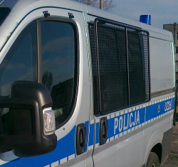 Zdjęcie kolorowe. Lewy bok radiowozu oznakowanego Fiat Ducato, koloru srebrnego z niebieską odblaskową taśmą przyklejoną w środkowej części pojazdu i napisem Policja.