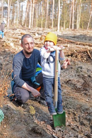 Zdjęcie kolorowe. Las. Kilkuletni chłopiec stoi na szpadlu wbitym w ziemie. Za nim przykuca mężczyzna.