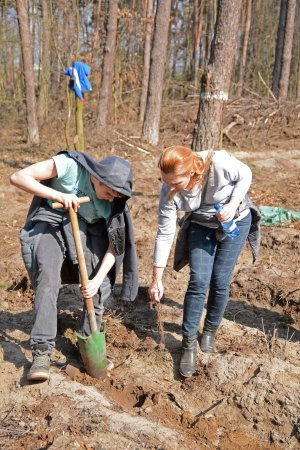 Zdjęcie kolorowe. Las. Z lewej strony młody chłopak wykopujący otwór w ziemi, z prawej pochylająca się kobieta, trzymająca w dłoni sadzonkę.