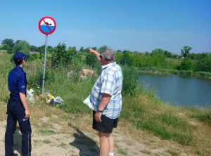 Zdjęcie kolorowe. Policjantka w umundurowaniu służbowym i stojący obok przedstawiciel WOPR sprawdzają oznakowanie niestrzeżonych kąpielisk.