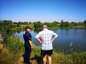 Zdjęcie kolorowe. Policjantka  oraz przedstawiciel WOPR stojący nad brzegiem akwenu wodnego, podczas wspólnej kontroli niestrzeżonych kąpielisk.