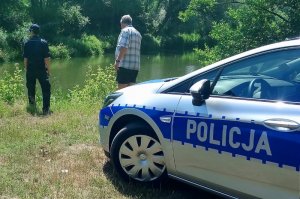 Zdjęcie kolorowe. Policjantka  oraz przedstawiciel WOPR stojący nad brzegiem akwenu wodnego, podczas wspólnej kontroli niestrzeżonych kąpielisk. Na pierwszym planie bok oznakowanego, policyjnego radiowozu.