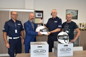 Zdjęcie kolorowe. Od lewej stoją kom. Artur Waszak Zastępca naczelnika Wydziału Ruchu Drogowego KMP w Kaliszu, Dyrektor Wojewódzkiego Ośrodka Ruchu Drogowego w Kaliszu Sławomir Chrzanowski, Komendant Miejski Policji w Kaliszu insp. Dariusz Bieniek oraz pracownik Wojewódzkiego Ośrodka Ruchu Drogowego w Kaliszu Radosław Paduch. Durektor WORD w trzyma w dłoniach kask motocyklowy koloru białego, który przekazuje Komendantowi Miejskiemu Policji w Kaliszu. Na stole stoją dwa kartony w który znajdowały się kaski. Na jednym z nich położony jest kask motocyklowy koloru białego.