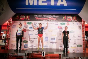 Zdjęcie kolorowe. Podium na którym stoją trzy laureatki III Kaliskiego Nocnego Półmaratonu „Bursztynowa Helena”. Kobieta stojąca na podium z nr 1 ma uniesione ręce do góry.