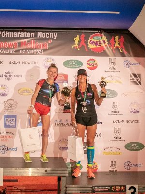 Zdjęcie kolorowe. Dwie kobiety uczestniczące w III Kaliskim Nocnym Półmaratonie „Bursztynowa Helena” stojące na podium z nr 1 i 3. W lewej dłoni trzymają puchary, w prawej torby koloru białego z nagrodami. Pozują fotografowi.