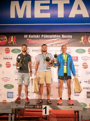 Zdjęcie kolorowe. Trzech mężczyzn uczestniczących w III Kaliskim Nocnym Półmaratonie „Bursztynowa Helena” stojących na podium z nr 1. W dłoniach trzymaka puchary oraz torby z nagrodami rzeczowymi. Pozują fotografom.