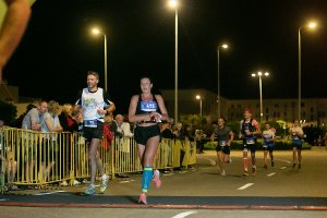 Zdjęcie kolorowe. Grupa biegaczy wbiegających na metę III Kaliskiego Nocnego Półmaratonu „Bursztynowa Helena”. Na pierwszym planie kobieta i mężczyzna za nimi grupa biegaczy. PO lewej stronie kibice stojący za barierkami ochronnymi.