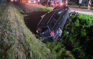 Zdjęcie kolorowe. Auto osobowe marki Opel, które wpadło do rowu po wcześniejszym zderzeniu z koparką. U góry zdjęcia widać jezdnię, na której zatrzymały się pojazdy i kierowców którzy wyszli ze swoich aut.