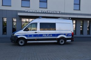 Zdjęcie kolorowe. Radiowóz oznakowany marki Volkswagen Crafter stojący przed głównym wejściem do Komendy Miejskiej Policja  w Kaliszu. W górnej części napis "Pomagamy i Chronimy".