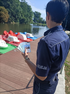 Zdjęcie kolorowe. Policjantka na przystani wodnej  w dłoni trzyma ulotki dotyczące bezpieczeństwa nad wodą.