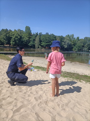 Policjantka na plaży przekazuje dzieciom lizaki, w drugiej dłoni trzyma ulotki dotyczące bezpieczeństwa nad wodą. W tle akwen wodny.