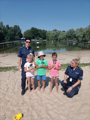 Zdjęcie kolorowe. Dwie policjantki na plaży z trójką dzieci pozują do zdjęcia.