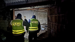 Zdjęcie kolorowe. Policjanci w umundurowaniu stający tyłem w pomieszczeniu, gdzie nocują bezdomni.