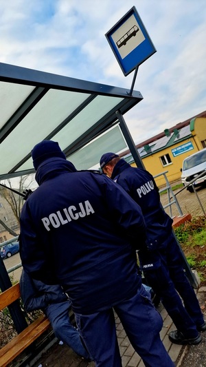 Zdjęcie kolorowe. Dwóch policjantów w umundurowaniu służbowym podczas rozmowy z osobami siedzącymi na przystanku autobusowym.