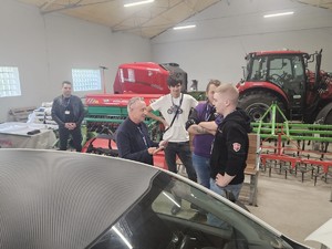 Zdjęcie kolorowe. Trzyosobowa drużyna uczniów szkoły średniej odpowiada na pytania egzaminatora  podczas eliminacji Ogólnopolskiego Młodzieżowego Turnieju Motoryzacyjnego. Na pierwszy planie fragment samochodu osobowego, w tle ciągnik i maszyny rolnicze.