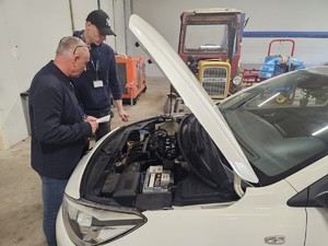 Zdjęcie kolorowe. Jeden z uczestników odpowiada na pytania egzaminatora  podczas eliminacji Ogólnopolskiego Młodzieżowego Turnieju Motoryzacyjnego. Uczeń i egzaminator stoją przy samochodzie osobowym, który ma otwartą pokrywę silnika.