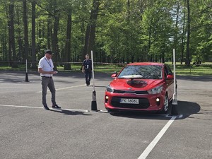 Zdjęcie kolorowe. Policjant i pracownik Wojewódzkiego Ośrodka Ruchu Drogowego w Kaliszu stojący na placu manewrowym obserwują uczestnika  podczas eliminacji Ogólnopolskiego Młodzieżowego Turnieju Motoryzacyjnego, który pokonuje plac samochodem osobowym.