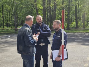 Zdjęcie kolorowe. Dwaj policjanci stojący na placu manewrowym rozmawiają ze stojącym obok mężczyzną.