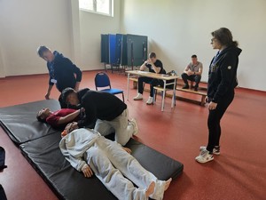 Zdjęcie kolorowe. Na sali, jedna z konkurencji - pierwsza pomoc przedmedyczna. Dwaj uczestnicy turnieju klęcząc na materacu udzielają pomocy osobą pokrzywdzonym. Jedna z osób stoi w pobliżu materaca, dwie inne siedzą w pobliżu.