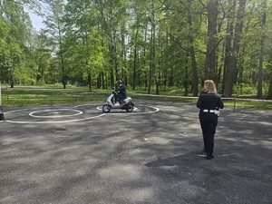 Zdjęcie kolorowe. Policjantka ruchu drogowego obserwuje uczestnika turnieju jadącego na motorowerze, który pokonuje przygotowany tor.