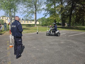 Zdjęcie kolorowe. Policjant ruchu drogowego obserwuje uczestnika turnieju jadącego na motorowerze, który pokonuje przygotowany tor.