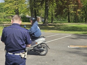 Zdjęcie kolorowe. Policjant ruchu drogowego obserwuje uczestnika turnieju jadącego na motorowerze, który pokonuje przygotowany tor.