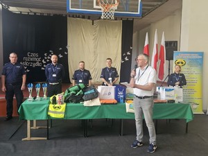 Zdjęcie kolorowe. Na pierwszy planie mężczyzna z mikrofonem odczytujący wyniki eliminacji Ogólnopolskiego Młodzieżowego Turnieju Motoryzacyjnego. W tle stół z nagrodami i pucharami oraz policjanci.