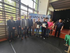 Zdjęcie Kolorowe. Trzyosobowa drużyna uczestników turnieju z opiekunem. Trzymają dyplomy i zdobyty puchar. Obok stoją przedstawiciele instytucji które organizowały turniej.