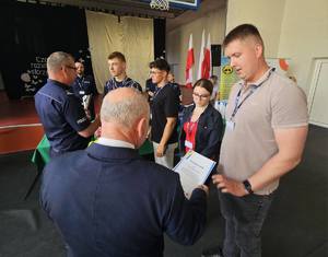 Zdjęcie Kolorowe. Komendant Miejski Policji w Kaliszu z drugim mężczyzną wręczają dyplom i puchar trzyosobowej drużynie uczestników turnieju oraz opiekunowi.